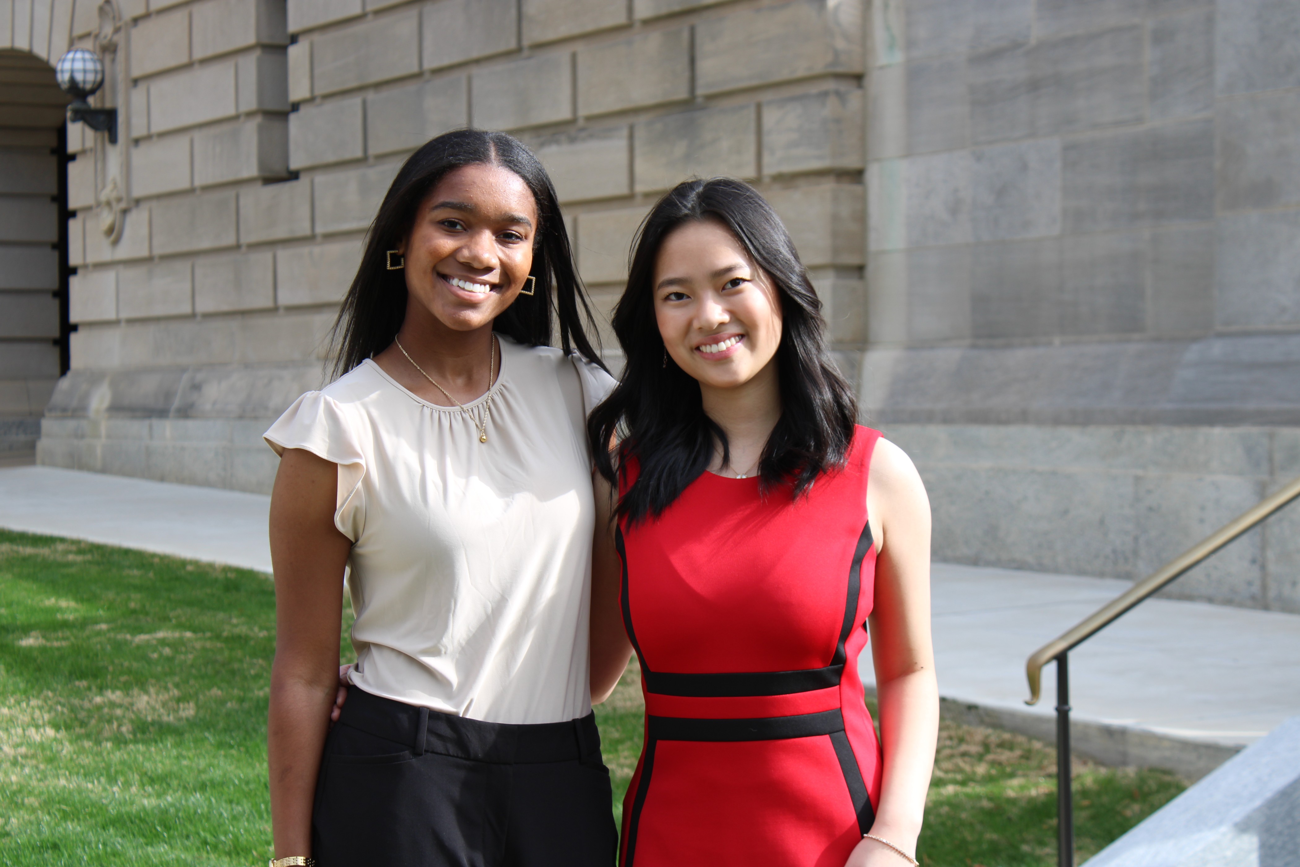 young African American female standing next to an Asian female outside of a brick building. While smiling and posing for a picture
